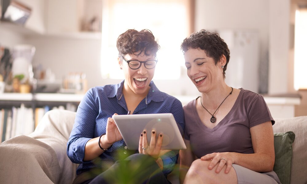 Zwei Frauen sitzen auf einem Sofa und schauen lachend auf ein Tablet. | © Getty Images / Tassii
