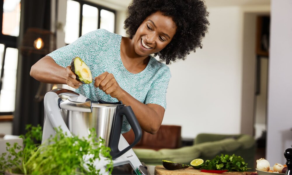Eine lächelnde Frau mit lockigem Haar gibt Avocado in einen Thermomix, während sie in einer hellen Küche steht. | © Vorwerk