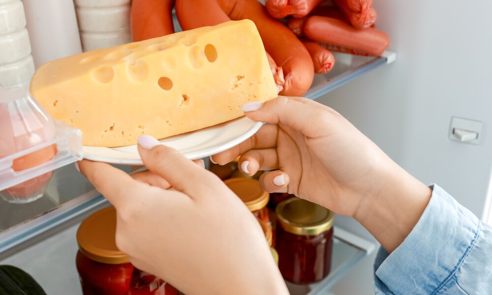 Zwei Hände legen ein Stück Emmentaler Käse auf einem Teller in einen offenen Kühlschrank voller Wurst und eingelegtem Gemüse. | © Adobe Stock/ Pixel-Shot