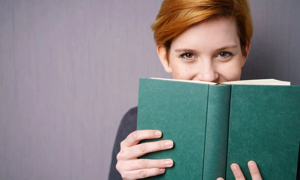 Lachende Frau versteckt sich hinter einem Buch | © adobe.stock.com/contrastwerkstatt