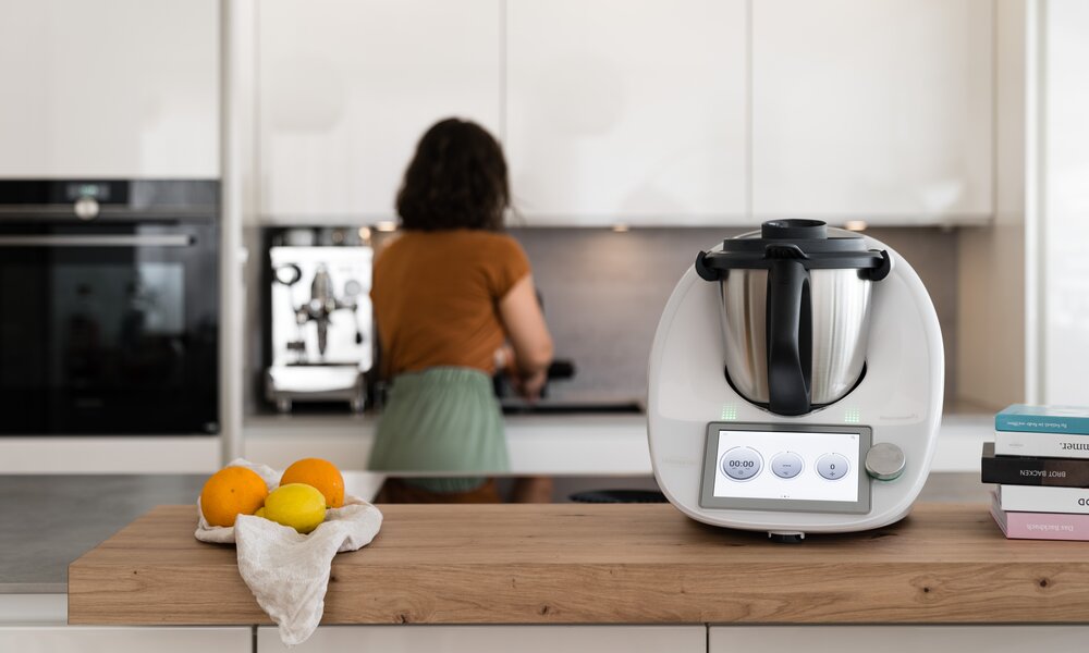 Thermomix TM6 auf der Küchenanrichte im Hintergrund Frau geblurrt am arbeiten. | © Vorwerk