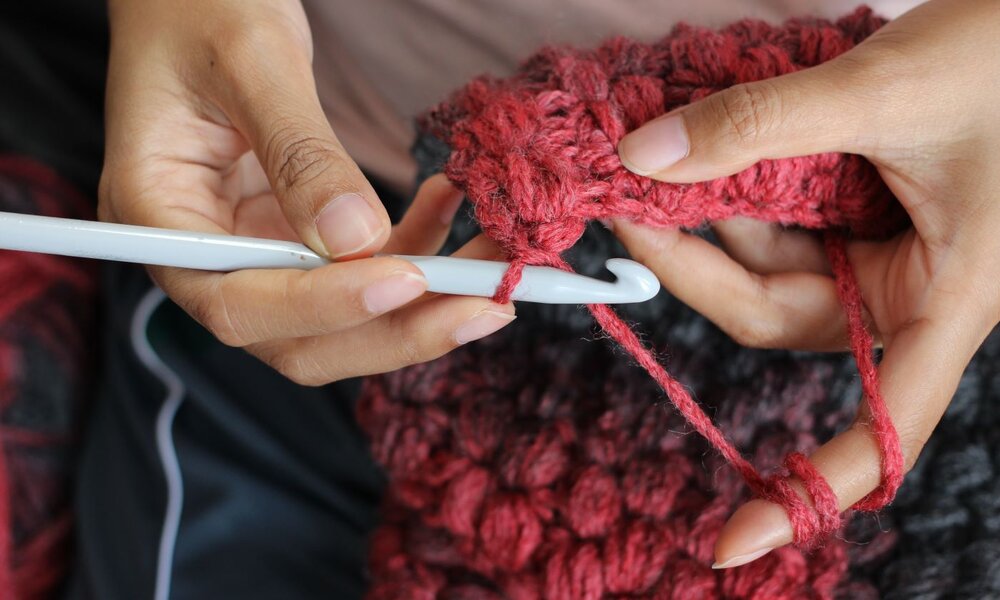 Nahaufnahme von Händen, die mit einer weißen Häkelnadel rotes Garn zu einem grob gestrickten Stück verarbeiten. | © nuankesaree/stock.adobe.com 