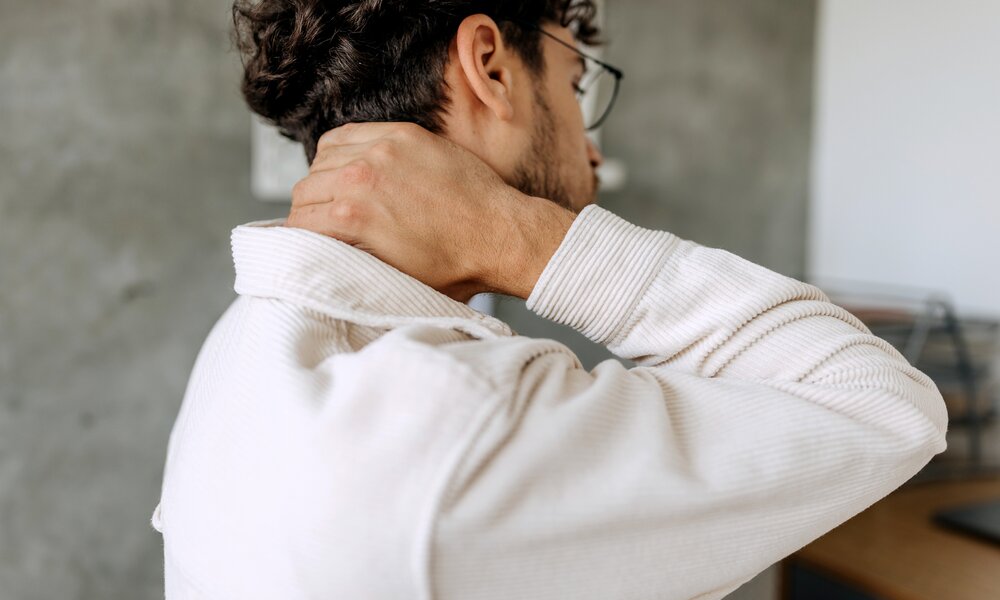 Seitenansicht eines Mannes mit gewelltem Haar und beigefarbener Jacke, der sich mit schmerzhaftem Ausdruck den Nacken massiert. | © Getty Images/Pekic