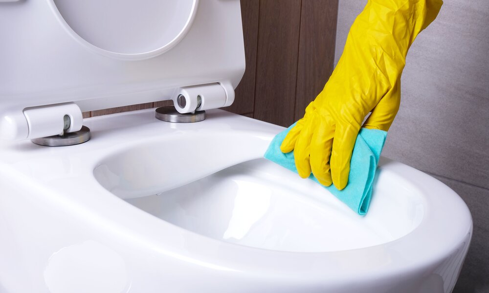 Eine Hand in gelbem Gummihandschuh reinigt mit einem blauen Tuch die weiße Toilettenschüssel in einem Badezimmer. | © Adobe Stock/ Alsu