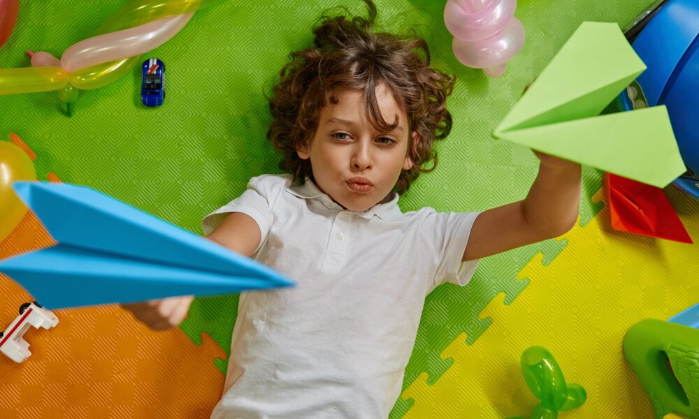 Ein Junge mit braunen Locken in einem weißen Polohemd liegt spielend auf bunten Matten und hält zwei Papierflieger in Blau und Grün. | © Nomad_Soul / stock.adobe.com