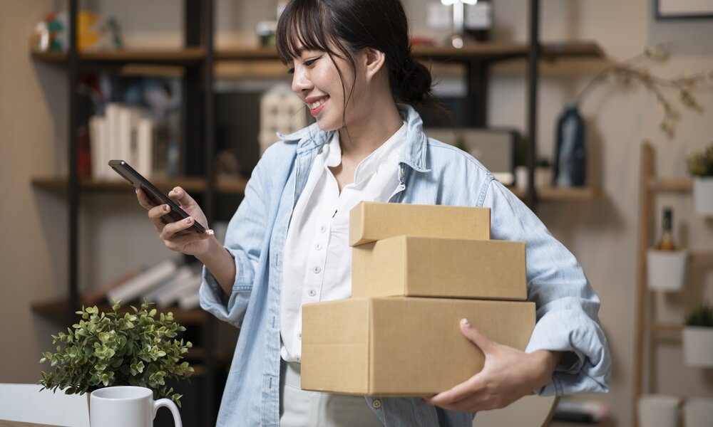 Eine junge Frau mit braunem Zopf und Jeanshemd lächelt, hält drei Kartons und tippt in einem hellen Arbeitszimmer auf ihr Smartphone. | © Getty Images/seksan Mongkhonkhamsao