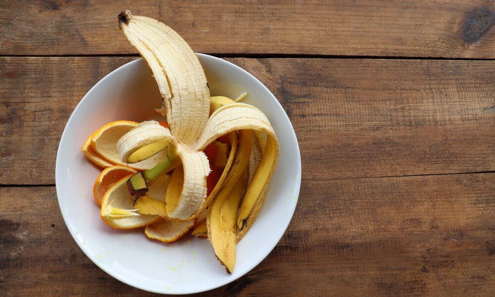 Eine geöffnete Bananenschale in einer weißen Schale liegt auf einem rustikalen Holztisch. | © Adobe Stock/ Juver