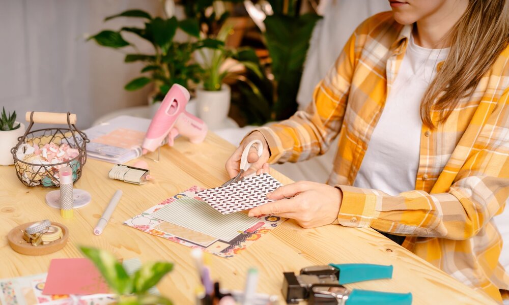 Eine Frau mit blondbraunen Haaren und kariertem Hemd bastelt an einem Holztisch mit Schere, Papier und Scrapbooking-Material. | © Anna/stock.adobe.com