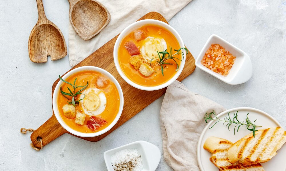 zwei Suppen in zwei weißen Schüssel stehen auf einem Holzbrett. Daneben befinden sich gegrillte Baguette-Scheiben und Holzbesteck. | © Getty Images / VictoriaBee