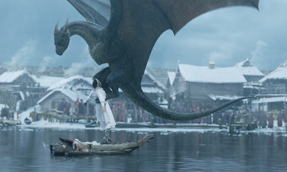 Vor winterlicher Kulisse ist ein Drache fast bis zur Oberfläche eines Sees hinuntergeflogen und packt mit seinen Klausen eine weißgekleidete junge Frau in einer Szene aus dem Kinofilm "Dragon: Love Is A Scary Tale" | © Amazon/Productbild zur DVD 