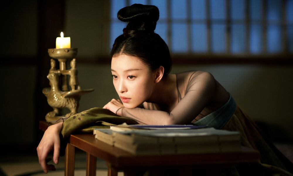 Schauspielerin Ni Ni stützt sich in einer Szene aus der Serie "The Rise of the Phoenixes" gedankenvoll auf einen niedrigen Tisch, ihr Gesicht erhellt von einer großen Kerze, die daneben steht. | © Netflix