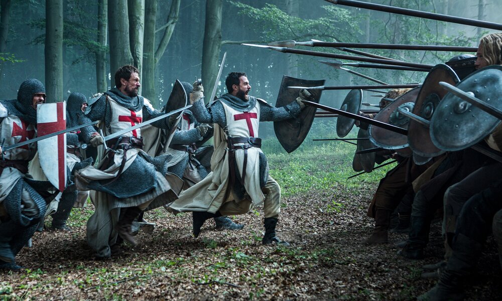 In einer Szene aus der Serie "Knightfall" rennen zwei verfeindete Rittertruppen in einem Wald mit erhobenen Waffen aufeinander zu. | © A+E Studios, LLC. All Rights Reserved.