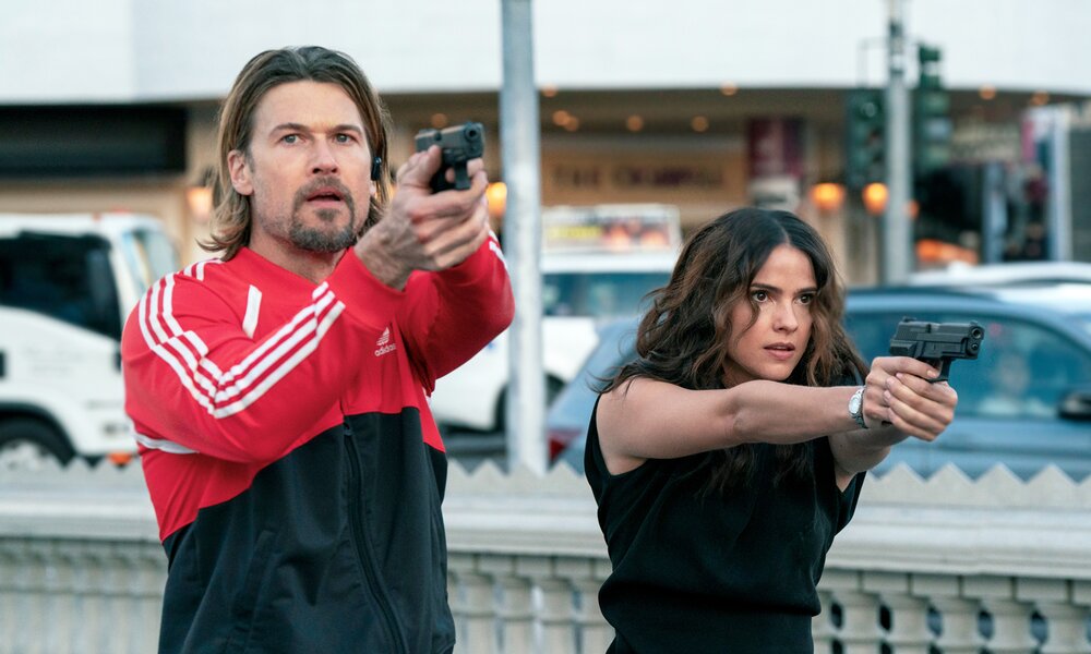 Nick Zano als Chad McKnight und Shelley Hennig als Ava Winters stehen in einer Szene der Netflix-Serie "Völlig zerstört" mit gezogenen Waffen auf der Straße. | © Netflix/iUrsula Coyote