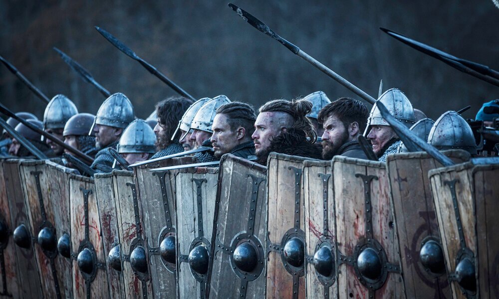  Arnas Fedaravicius als Sihtric, Alexander Dreymon als Uhtred und Mark Rowley als Finan imit anderen Kämpfern in Rüstung stehen in einer Szene aus dem Netflix-Film "Seven Kings Must Die" kampfbereit nebeneinander hinter ihren aufgestellten Schilden. | © Netflix/&/