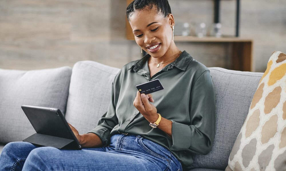 Frau in grüner Bluse und blauer Jeans sitzt lächelnd auf einem Sofa und hält eine Kreditkarte in der Hand, während sie ein Tablet benutzt. | © Getty Images/Charday Penn