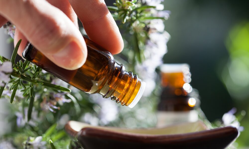 Nahaufnahme einer Hand, die ätherisches Öl aus einer braunen Flasche in eine Keramikschale gießt, im Hintergrund Rosmarinzweige. | © Adobe Stock/ plprod