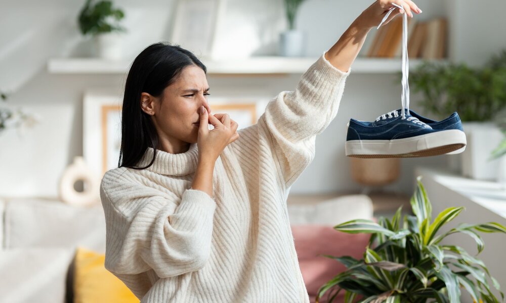 Eine junge Frau in beigem Pullover hält eine blaue Turnschuh mit angewidertem Gesichtsausdruck und hält sich die Nase zu. | © Adobe Stock/ nenetus