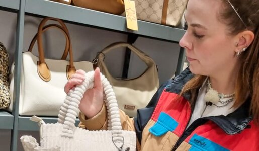 Junge Frau mit braunem Haar und Haarreif betrachtet eine cremefarbene Handtasche in einem Laden, umgeben von weiteren Taschen. | © RTL/Constantin Ent.