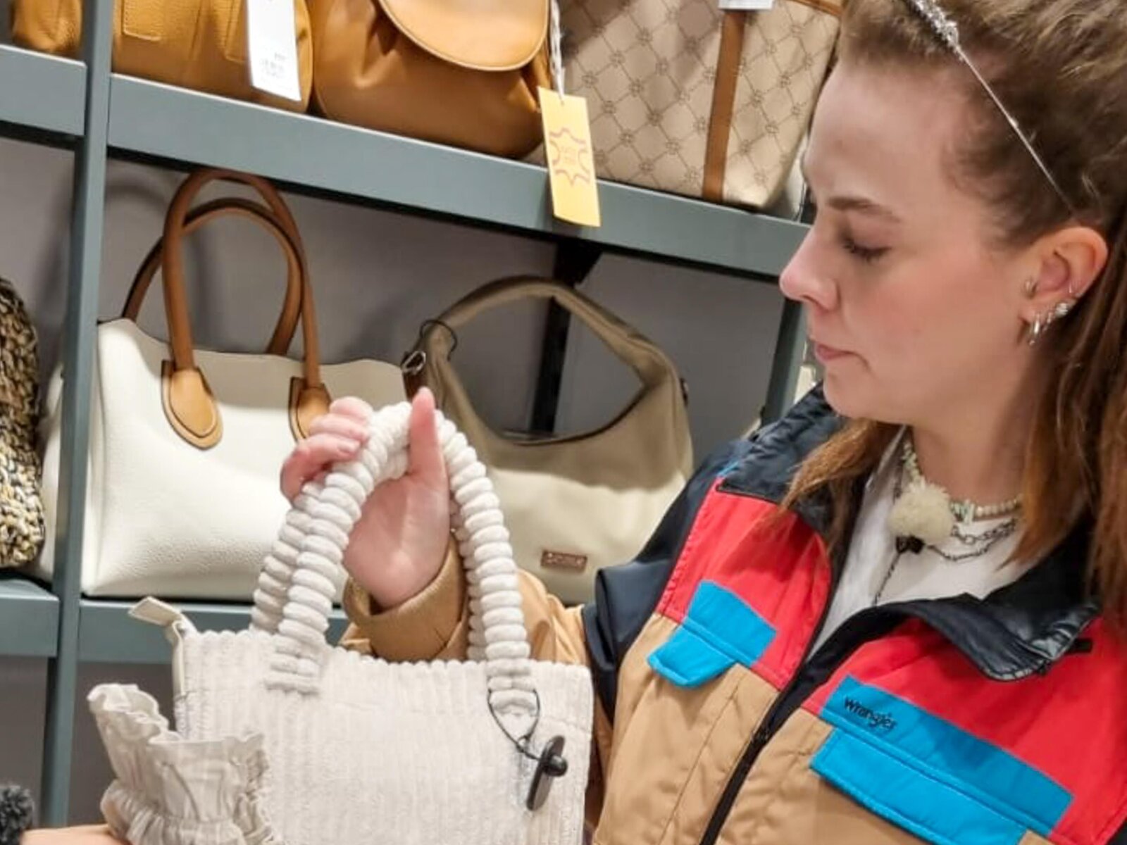 Junge Frau mit braunem Haar und Haarreif betrachtet eine cremefarbene Handtasche in einem Laden, umgeben von weiteren Taschen. | © RTL/Constantin Ent.