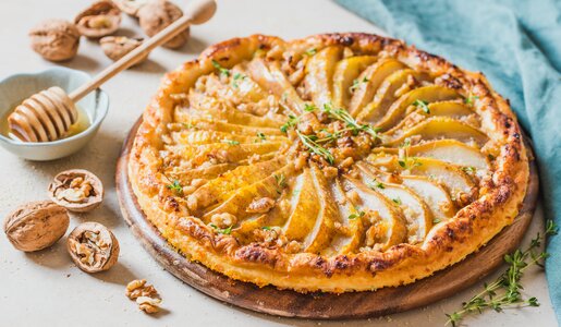 Birnenkuchen mit sauber drapierten Birnenscheiben, darüber glasierte Walnüsse und daneben steht eine kleine Schale mit Honig. | © Adobe Stock / travelbook
