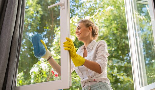 Junge blonde Frau in gelbem Gummihandschuh reinigt mit einem blauen Schwamm lächelnd ein geöffnetes Fenster bei Sonnenschein. | © Adobe Stock/ cherryandbees