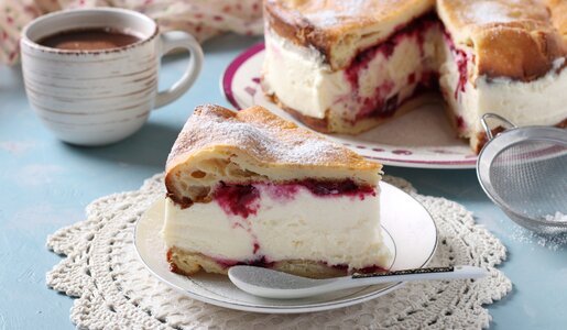 Stück Joghurtkuchen mit Kirschen auf einem Teller, dahinter der angeschnittene, ganze Kuchen. | © Adobe Stock / Anzhela
