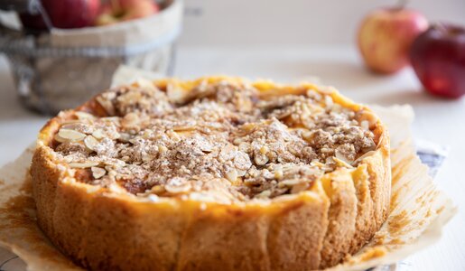Apfelkuchen auf einem Kuchengitter, darauf Backpapier und im Hintergrund unscharf Äpfel. | © Adobe Stock / Kerstin