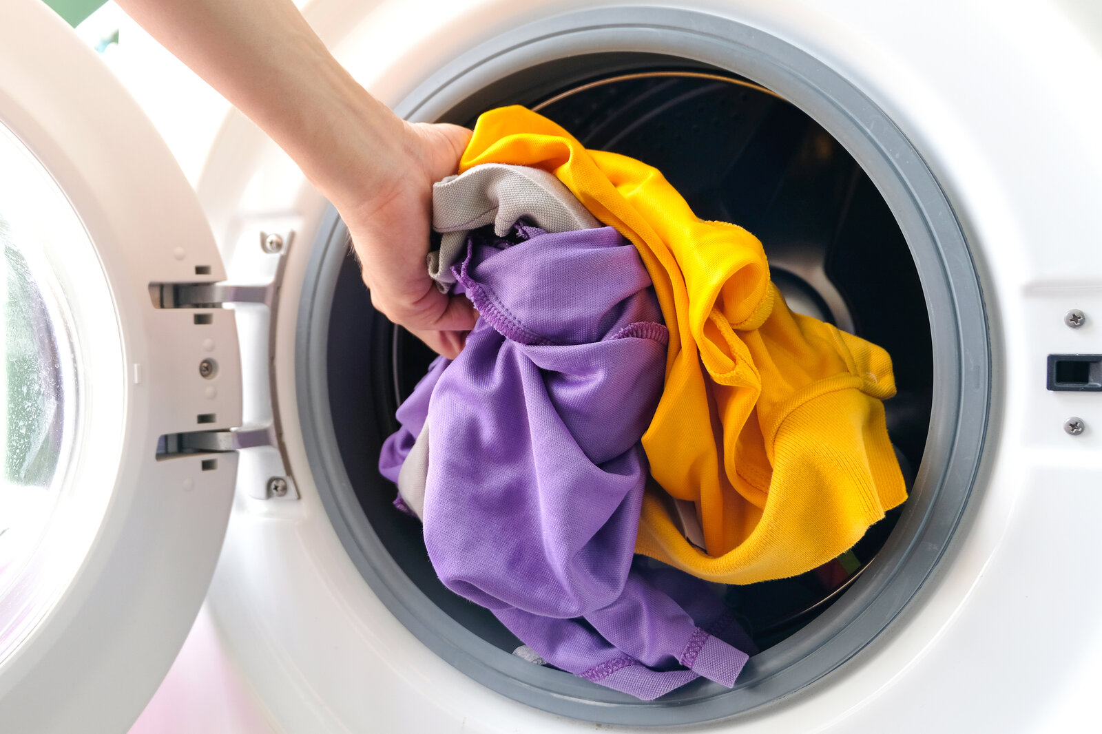 Eine Hand legt bunte Wäsche in Gelb, Lila und Grau in die geöffnete Trommel eines Wäschetrockners | © Adobe Stock/ Photo Sesaon