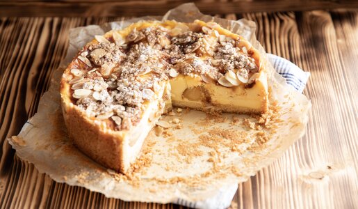 Angeschnittener Apfel-Schmandkuchen auf etwas Backpapier und einem Küchentuch angerichtet, auf einem Holzuntergrund stehend. | © Adobe Stock / Kerstin
