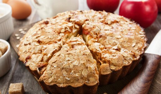 Angeschnittener Apfel-Haferflockenkuchen mit frischen Äpfeln, Eiern und Haferflocken im Hintergrund. | © Adobe Stock /  minadezhda