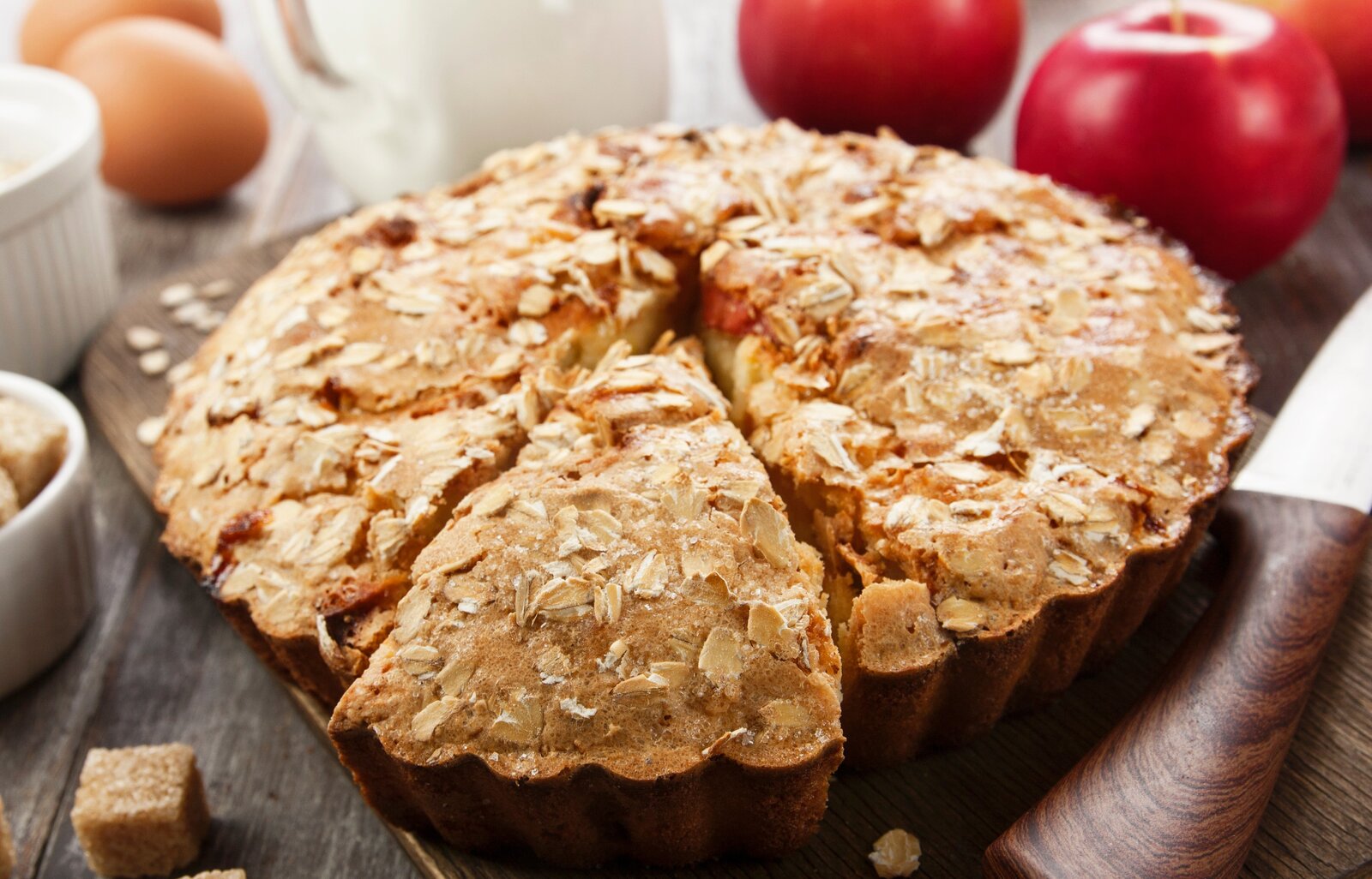 Angeschnittener Apfel-Haferflockenkuchen mit frischen Äpfeln, Eiern und Haferflocken im Hintergrund. | © Adobe Stock /  minadezhda