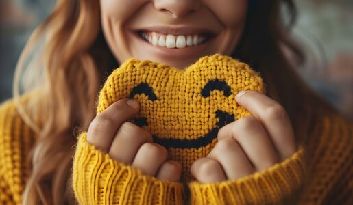 Frau in gelbem Strickpullover hält ein gehäkeltes Smiley-Gesicht und lächelt breit, Nahaufnahme ihres Gesichts. | © Yiss Design/stock.adobe.com
