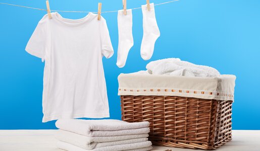 Ein weißes T-Shirt und Socken hängen an einer Wäscheleine vor blauem Hintergrund, daneben ein Korb mit Handtüchern. | © Adobe Stock/ LIGHTFIELD STUDIOS
