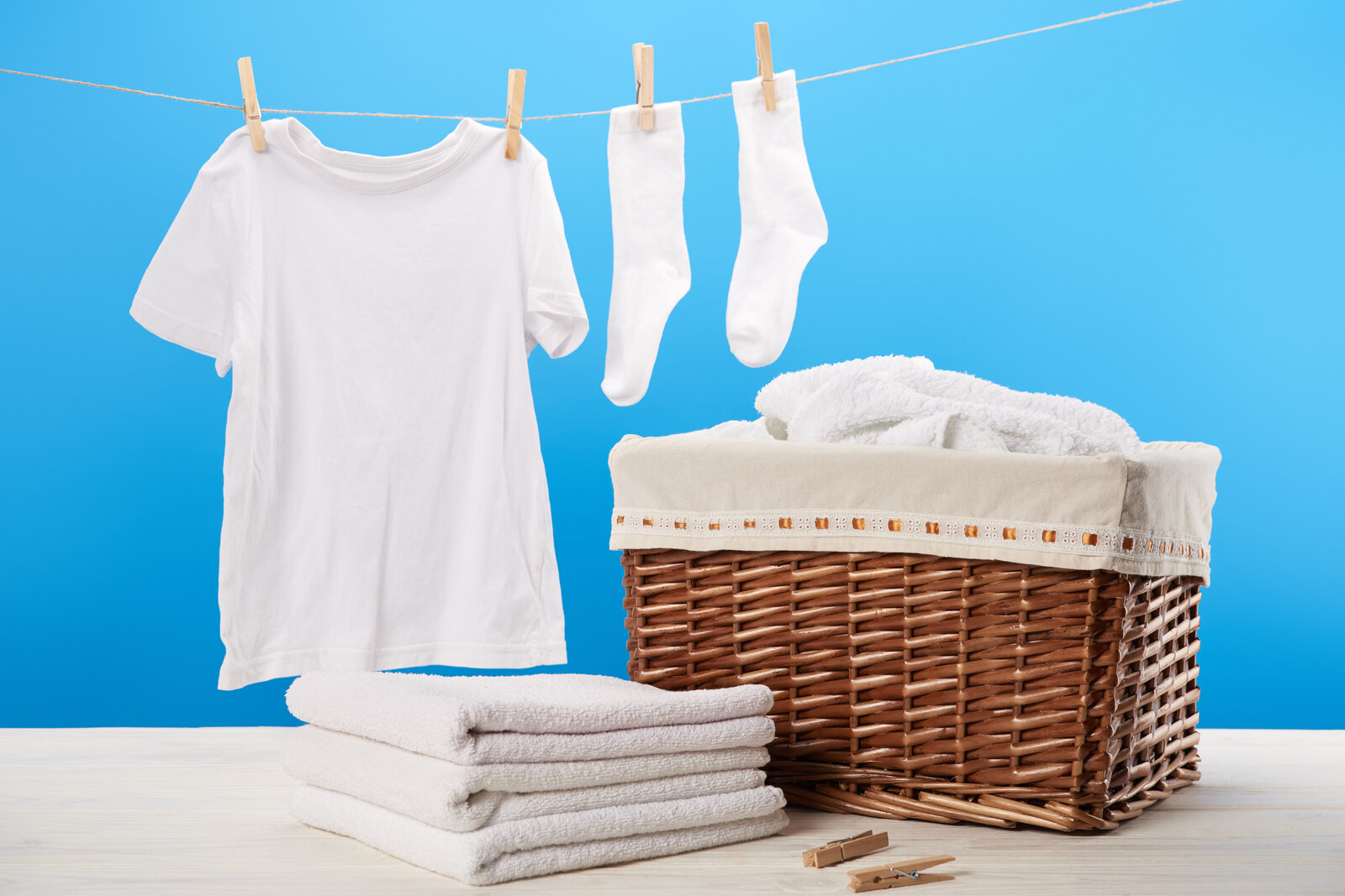 Ein weißes T-Shirt und Socken hängen an einer Wäscheleine vor blauem Hintergrund, daneben ein Korb mit Handtüchern. | © Adobe Stock/ LIGHTFIELD STUDIOS