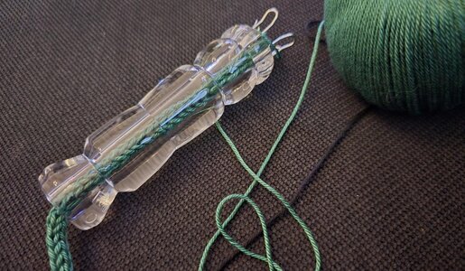 Eine durchsichtige Strickliesel mit grüner Wolle liegt auf einem dunklen, strukturierten Stoffhintergrund. | © Sabrina Forst