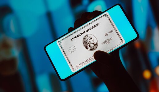 Smartphone mit leuchtendem Bildschirm zeigt silberfarbene American Express Kreditkarte, dahinter verschwommener Hintergrund in Blautönen. | © IMAGO/Zoonar