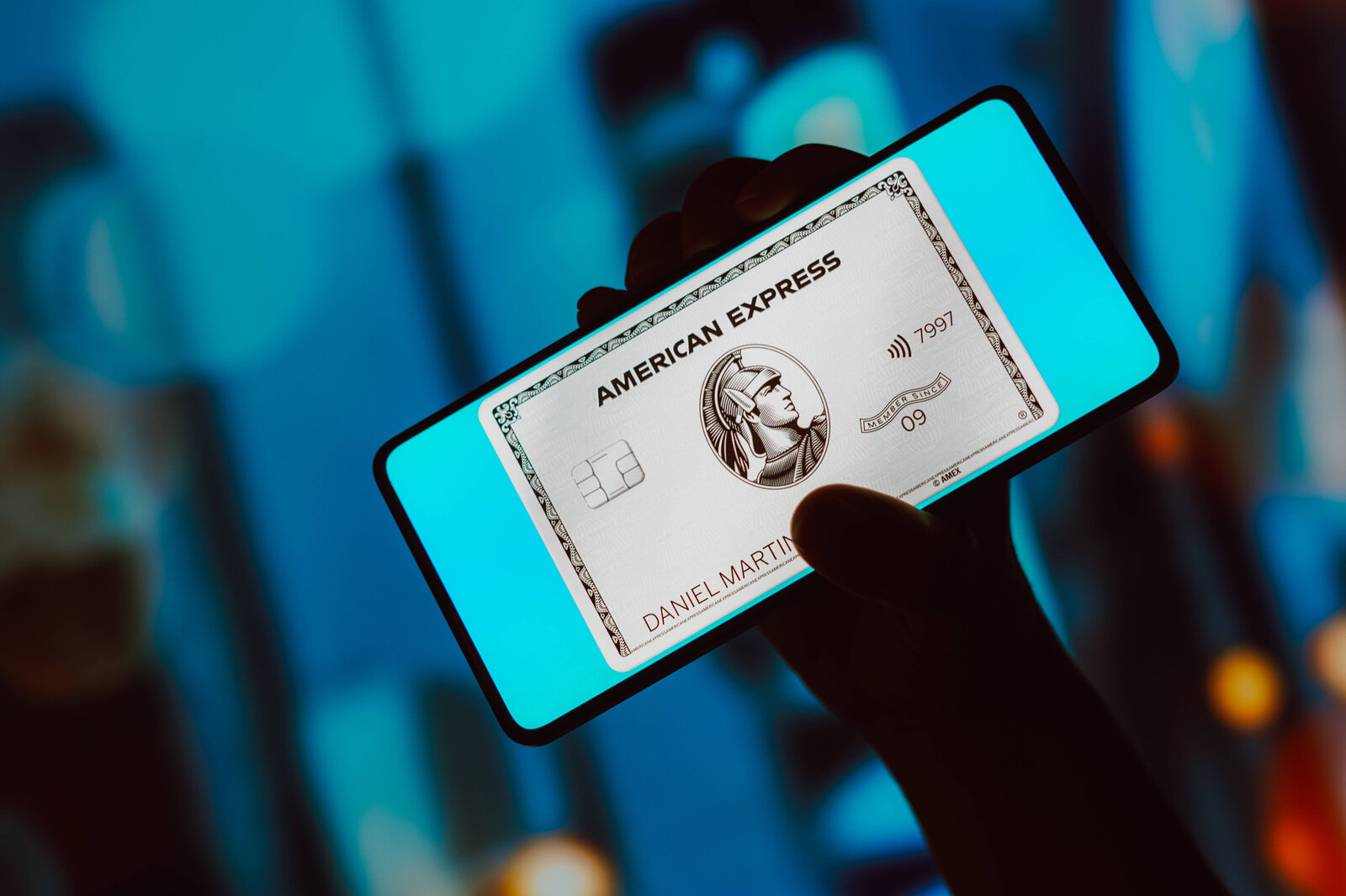 Smartphone mit leuchtendem Bildschirm zeigt silberfarbene American Express Kreditkarte, dahinter verschwommener Hintergrund in Blautönen. | © IMAGO/Zoonar