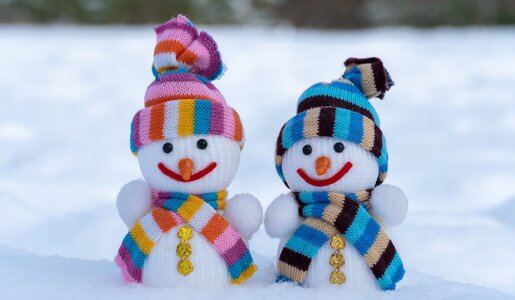 Zwei lächelnde Schneemänner mit bunter Strickmütze und Schal stehen nebeneinander auf schneebedecktem Boden vor verschwommenem Hintergrund. | © IhorStore / stock.adobe.com