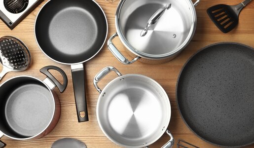 Ein Set aus Edelstahl-Töpfen und Pfannen mit schwarzen Griffen liegt auf einer Holzarbeitsplatte, umgeben von Küchenutensilien. | © Adobe Stock/ New Africa