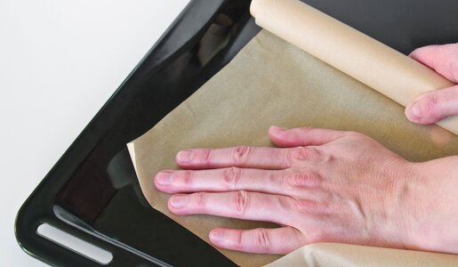 Backpapier auf schwarzem Backblech bildfüllend, Hand streicht Backpapier glatt auf das Blech | © Adobe Stock/Gehkah