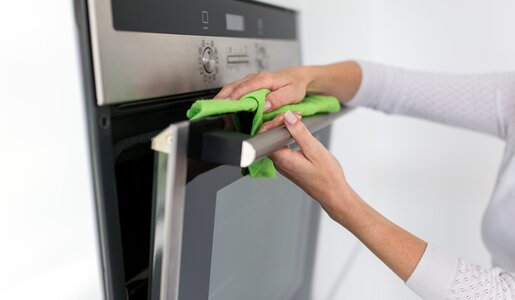 Silberner Backofen bildfüllend, Hände einer Frau sind am Backofen und putzen sie mit einem grünen Tuch sauber | © Adobe Stock/mmphotographie.de