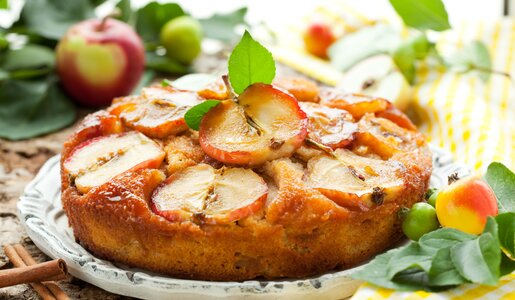 Apfelkuchen belegt mit Apfelscheiben auf einem weißen Teller, im Hintergrund frische Äpfel und Blätter. | © Adobe Stock / Svetlana Kolpakova