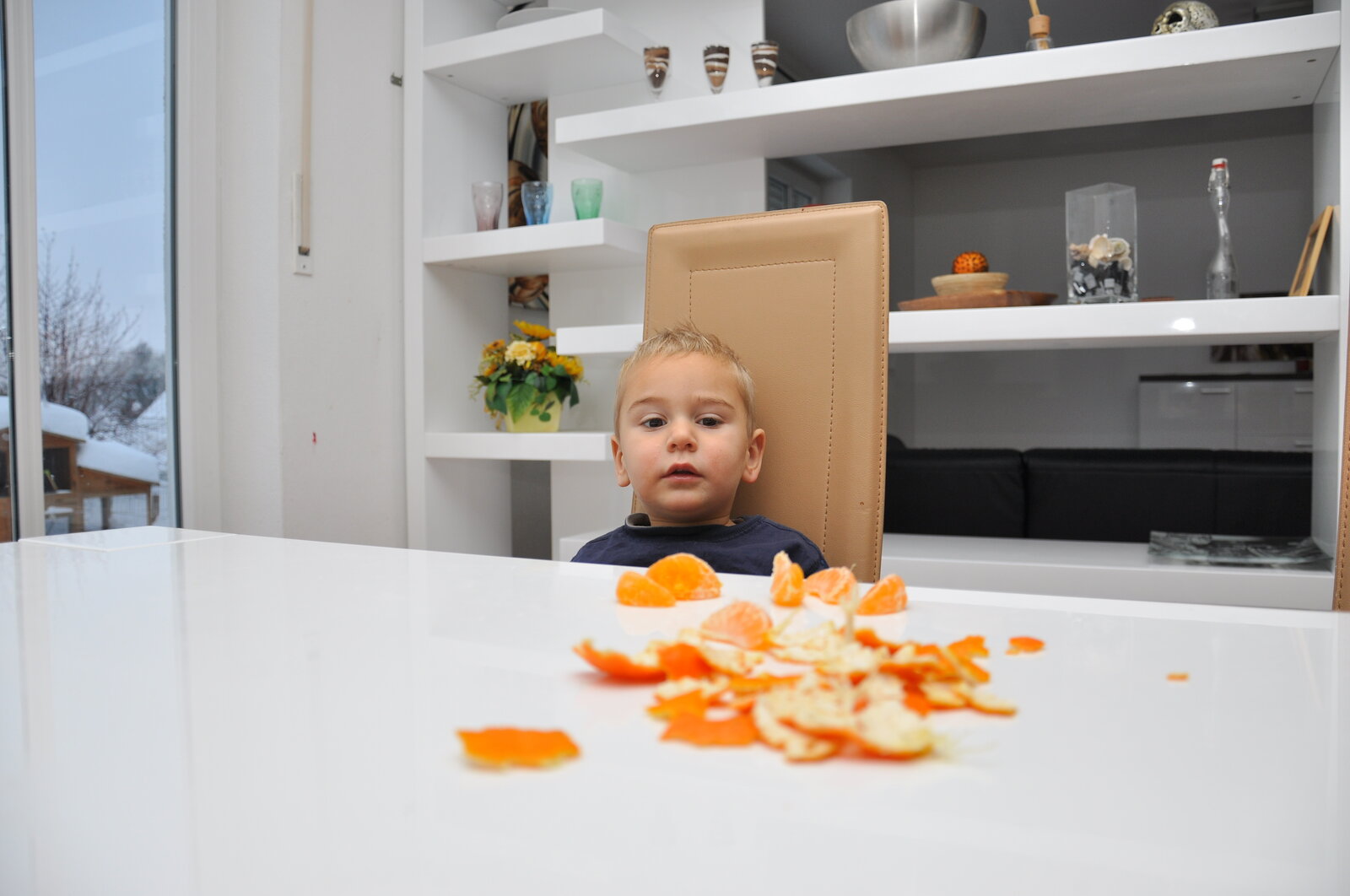 Großer weißer Tisch in der unteren Hälfte des Bildes, darauf liegen Mandarinenschalen und eine Mandarine in Orange, vor dem Tisch sitzt ein kleiner Junge in blau gekleidet und guckt die Mandarinen an, dahinter ein Regal | © Adobe Stock/Bernd Libbach
