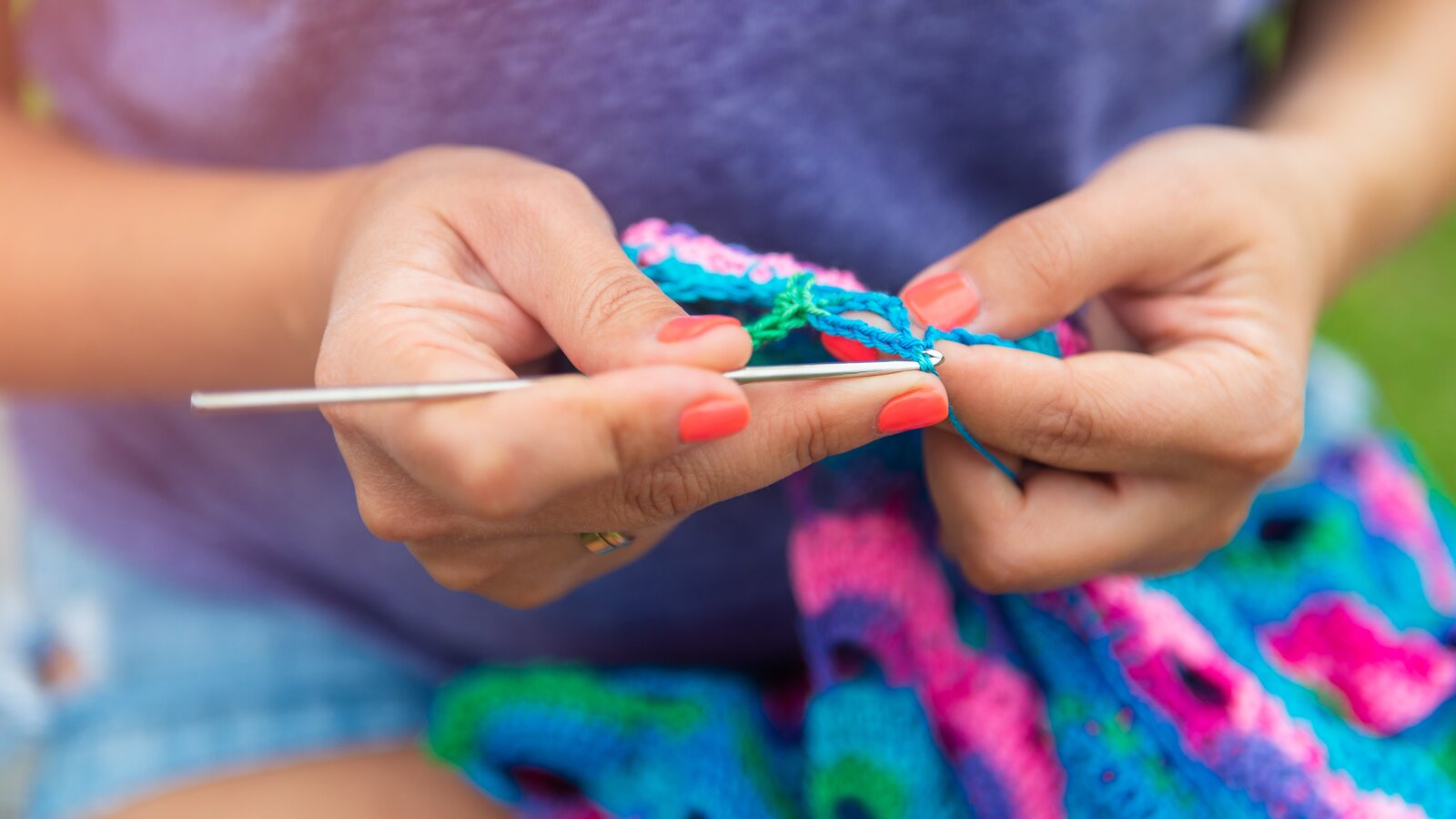 Eine Frau in lila Shirt häkelt ein buntes Tuch in kräftigen Farben, die Hände präzise und konzentriert im Fokus. | © manuta/ stock.adobe.com