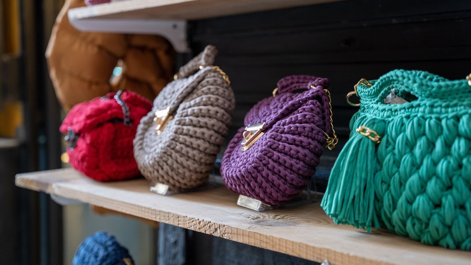 Vier handgefertigte Häkelhandtaschen in Rot, Beige, Lila und Grün mit goldenen Kettenhenkeln stehen auf einem Holzregal. | © Erman Gunes / stock.adobe.com