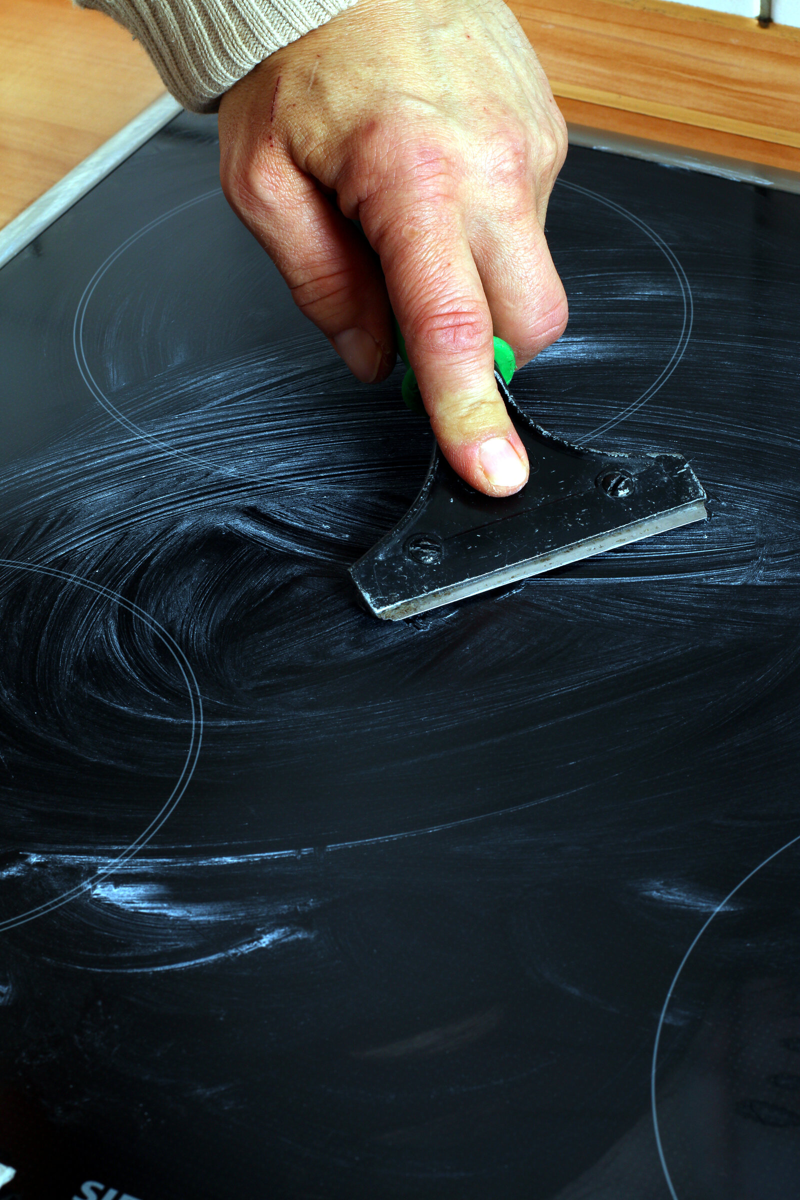 Glaskeramikkochfeld in schwarz, eine männliche Hand scharbt mit Scharber ein weißes Reinigungsmittel darüber  | © Adobe Stock/Osterland