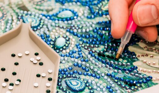 Eine beige Schale mit glitzernden Strasssteinen neben einer Hand, die ein detailreiches Kunstwerk aus Schmucksteinen gestaltet. | © iStock / Getty Images Plus / Daria Nipot