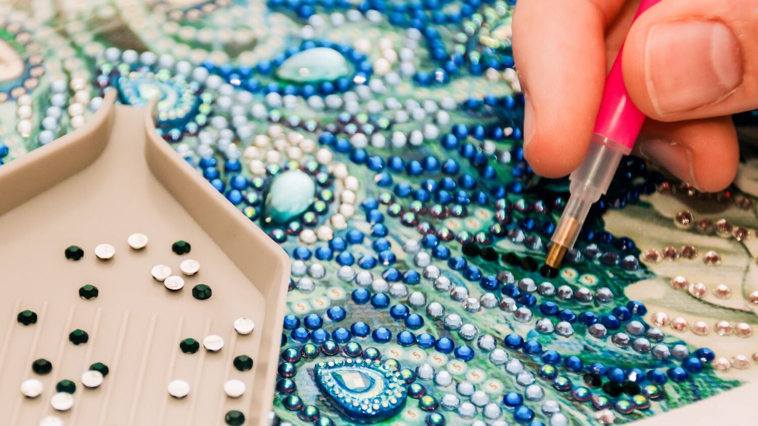 Eine beige Schale mit glitzernden Strasssteinen neben einer Hand, die ein detailreiches Kunstwerk aus Schmucksteinen gestaltet. | © iStock / Getty Images Plus / Daria Nipot