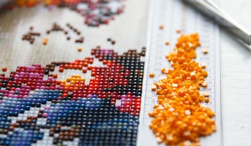 Nahaufnahme eines halbfertigen Diamond-Painting-Motivs mit farbigen Steinen, gelben Perlen und Pinzette auf einem Raster. | © Mariana Rusanovschi /stock.adobe.com