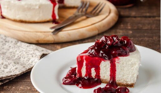 Stück Käsekuchen mit Kirschkompott auf einem Teller, dahinter der gesamte Kuchen. | © Adobe Stock / yuliiaholovchenko